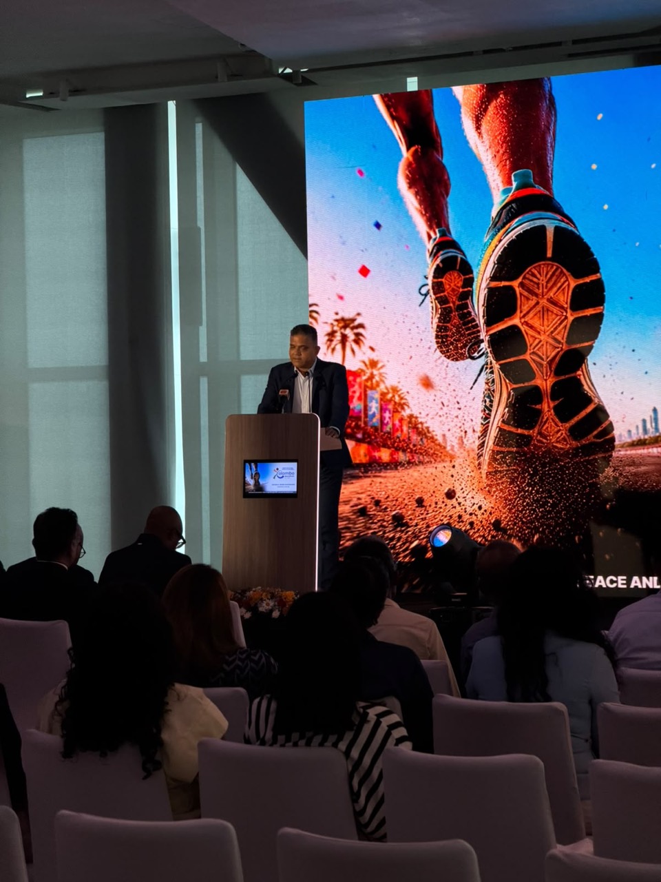 Speaker at a lectern beside an LED screen showing a runner's shoes